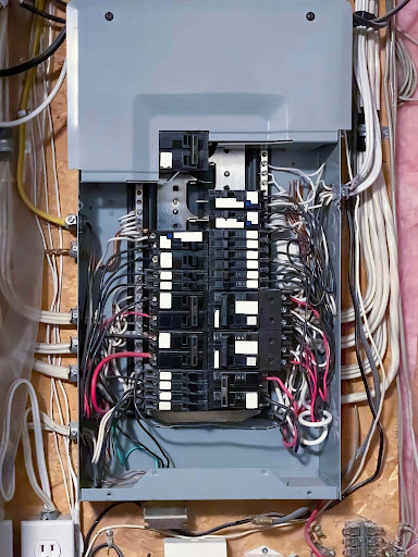 The image shows an open circuit breaker box. The breakers and wires are exposed.